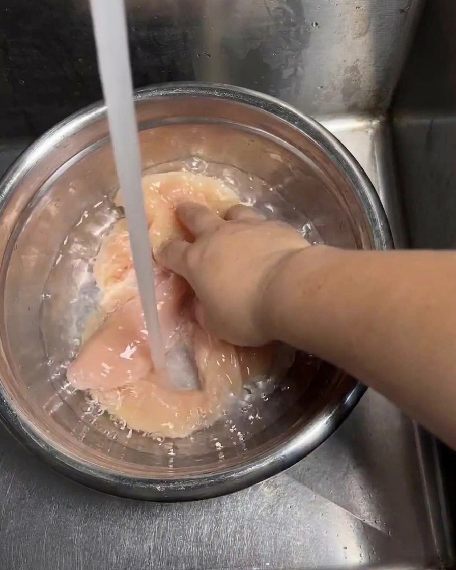 Un chorro de agua fría del grifo llenando un tazón de acero inoxidable con filetes de pollo crudos mientras una mano los mueve para enjuagarlos.