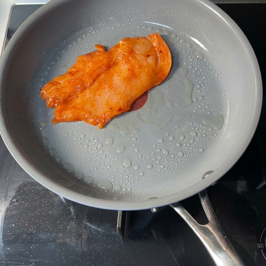Un filete de pollo marinado de color naranja cocinándose en una sartén antiadherente gris con gotas visibles de aceite en la superficie.