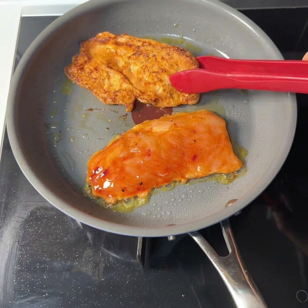 Unas pinzas rojas volteando un filete de pollo marinado en una sartén gris, mostrando una costra dorada y sellada en el lado cocido.