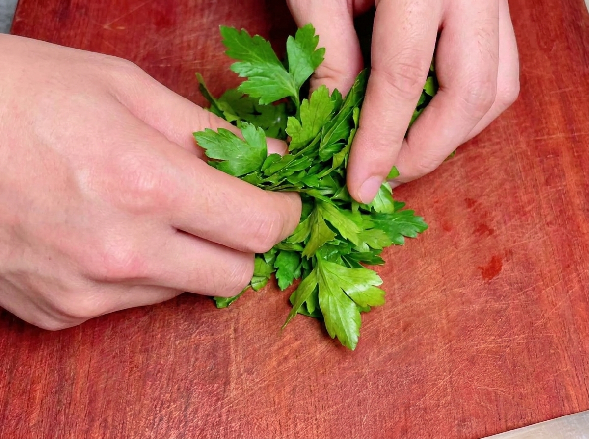Dos manos rolling up a bunch of fresh green flat-leaf parsley tightly on a red-brown wooden cutting board.