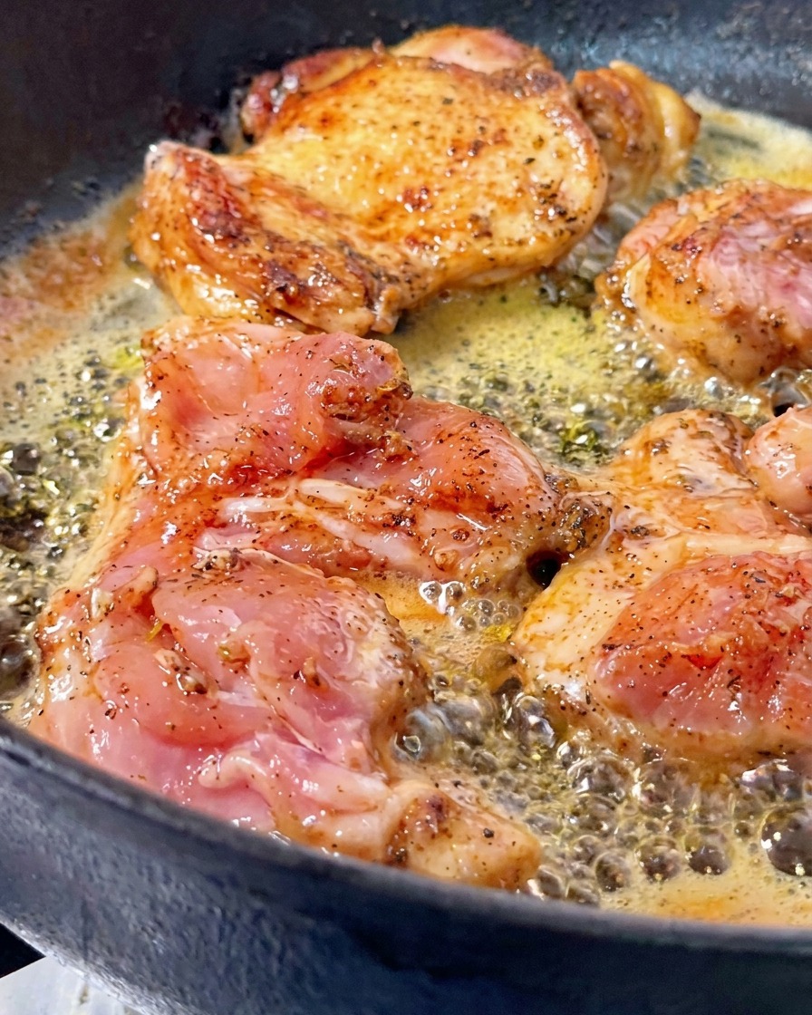 Contramuslos de pollo dorándose en mantequilla burbujeante dentro de una sartén de hierro fundido, con una pieza volteada que muestra una costra crujiente de color marrón dorado.
