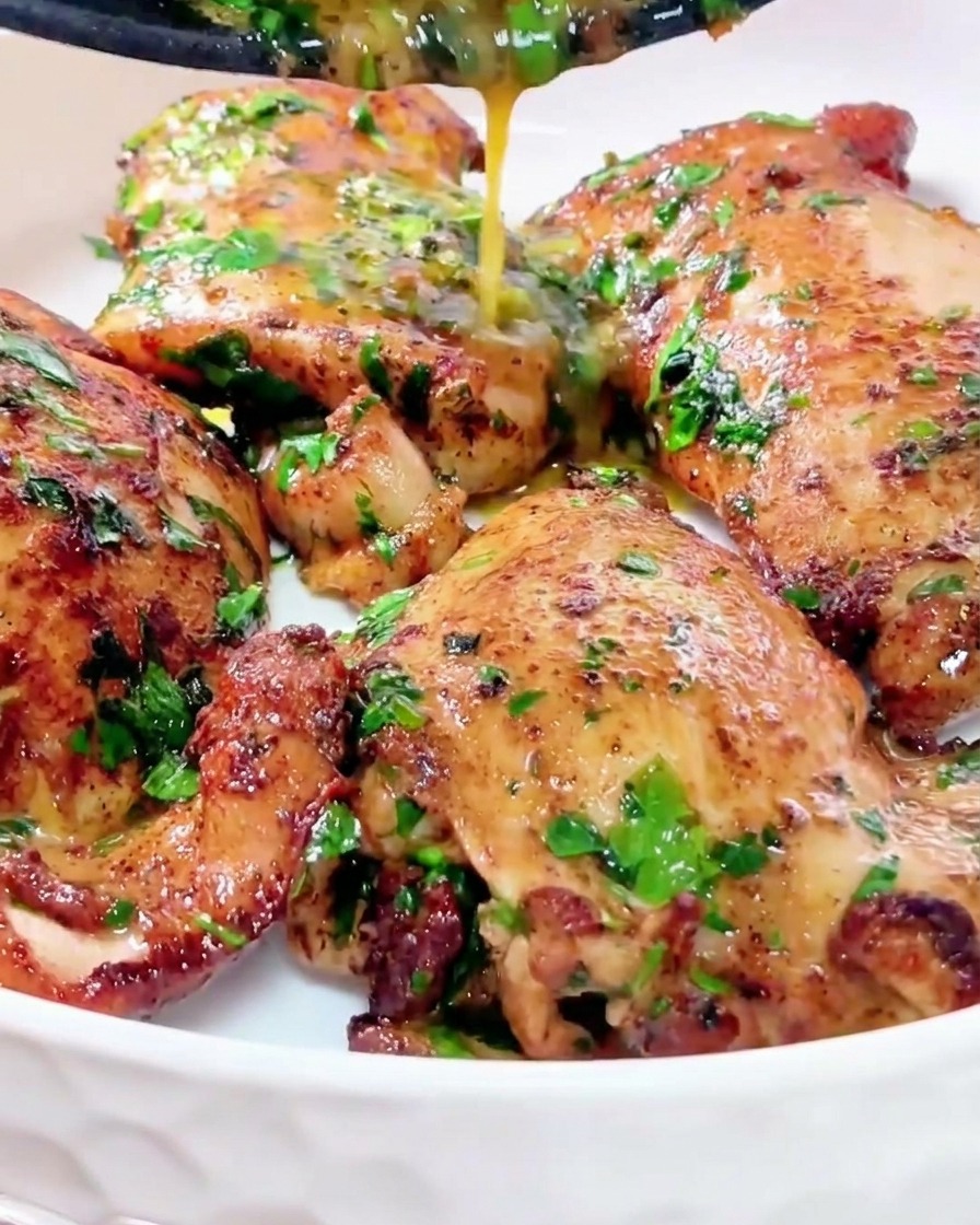 Un hilo de salsa de mantequilla dorada y brillante mezclada con hierbas verdes cayendo desde una sartén sobre jugosos contramuslos de pollo de color marrón dorado en un bol blanco limpio.