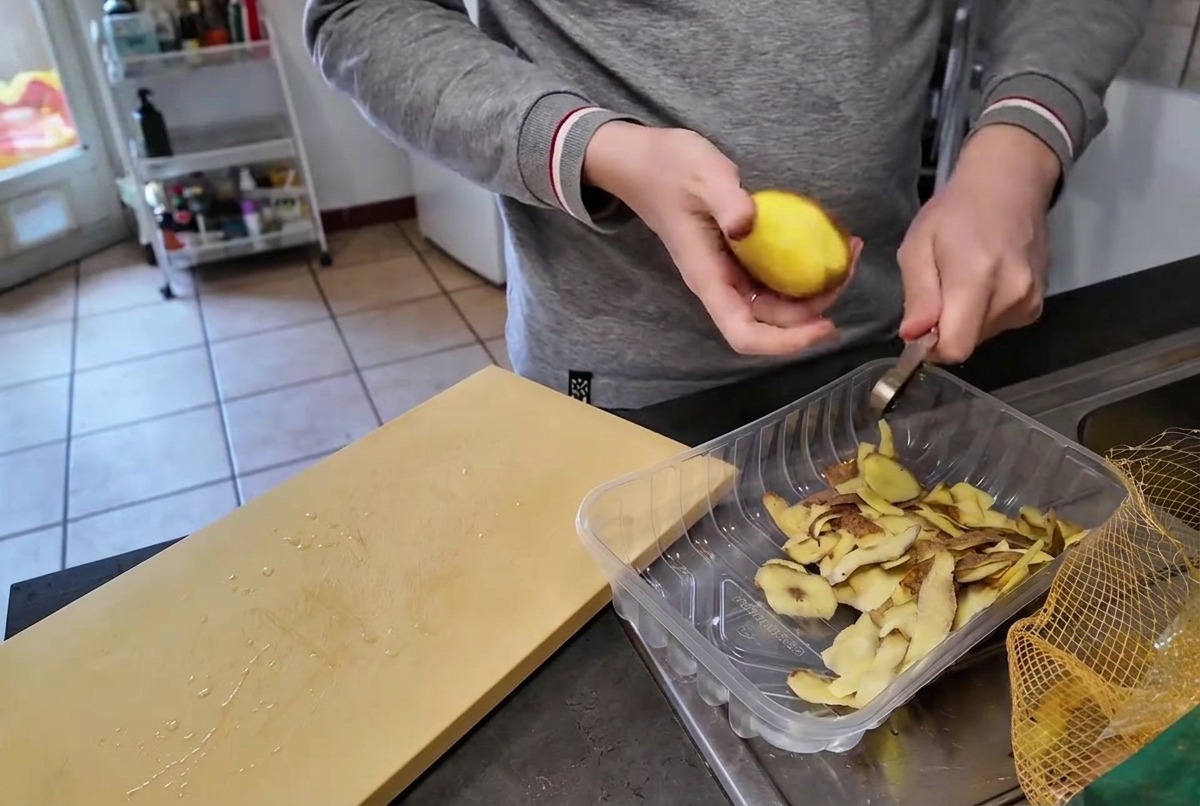 Una persona pelando una patata amarilla cruda con un cuchillo pequeño sobre un recipiente transparente que contiene las mondas.