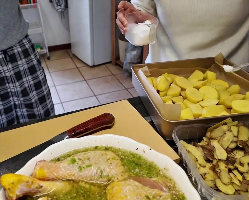 Trozos de patata cruda distribuidos en una bandeja de horno forrada con papel vegetal, siendo sazonados con sal desde un recipiente.