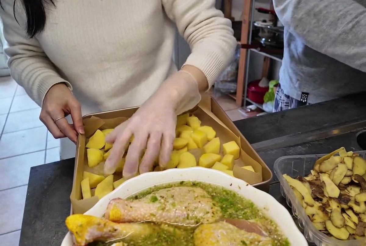 Una mano con un guante de plástico transparente mezclando los trozos de patata en una bandeja de horno de metal.