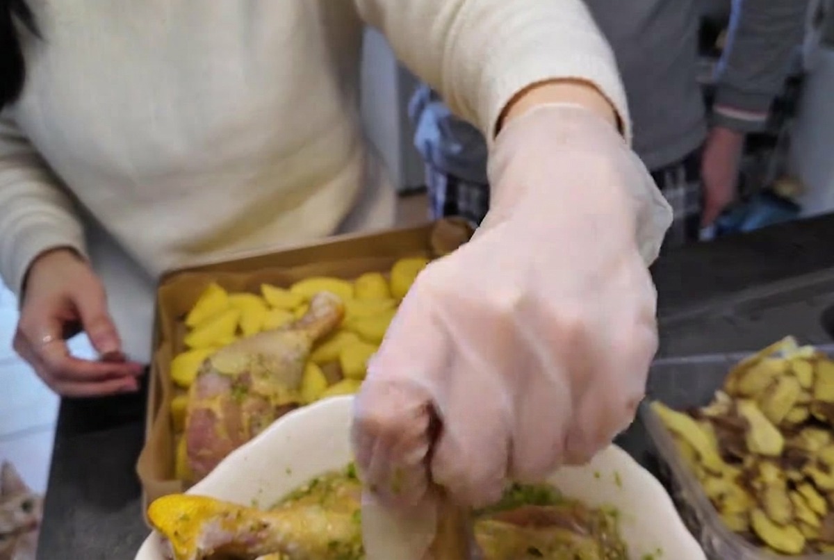Una mano con guante levantando un muslo de pollo crudo cubierto de marinada de hierbas verdes de un bol blanco.