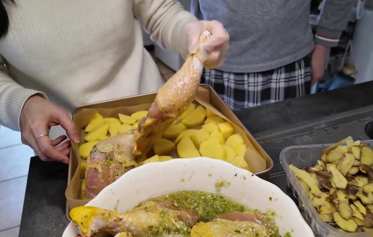 Muslos de pollo crudos marinados siendo colocados con la piel hacia arriba sobre una capa de patatas troceadas en una bandeja de horno dorada.