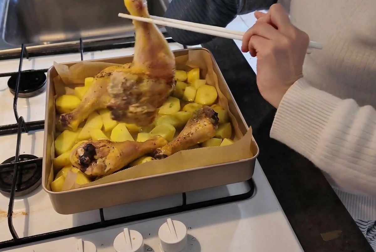 Una persona con una manopla de horno usando pinzas o palillos para dar la vuelta a un muslo de pollo a medio hacer en una bandeja caliente con patatas.