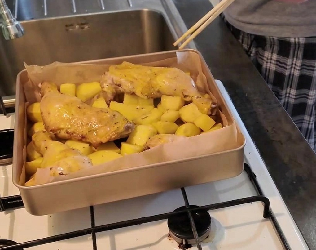 Una persona usando unos palillos para pinchar la carne de un muslo de pollo sobre una cama de patatas y comprobar si está hecho.