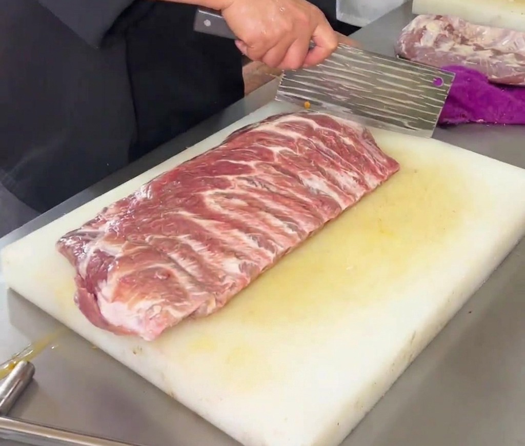 Un chef usando un gran hacha de cocina para marcar un patrón de diamante en un costillar crudo de cerdo sobre una tabla de cortar de plástico blanco.