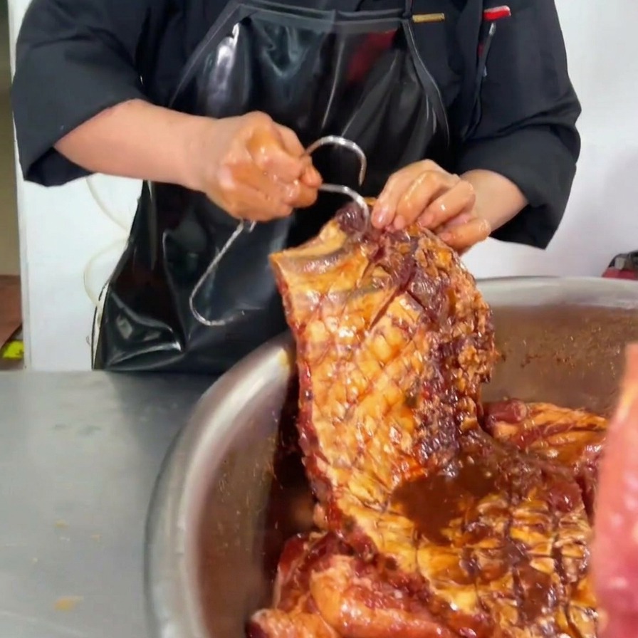Un chef insertando un gran gancho de metal en la esquina superior de un costillar de cerdo completamente marinado con salsa oscura sobre un tazón de acero inoxidable.