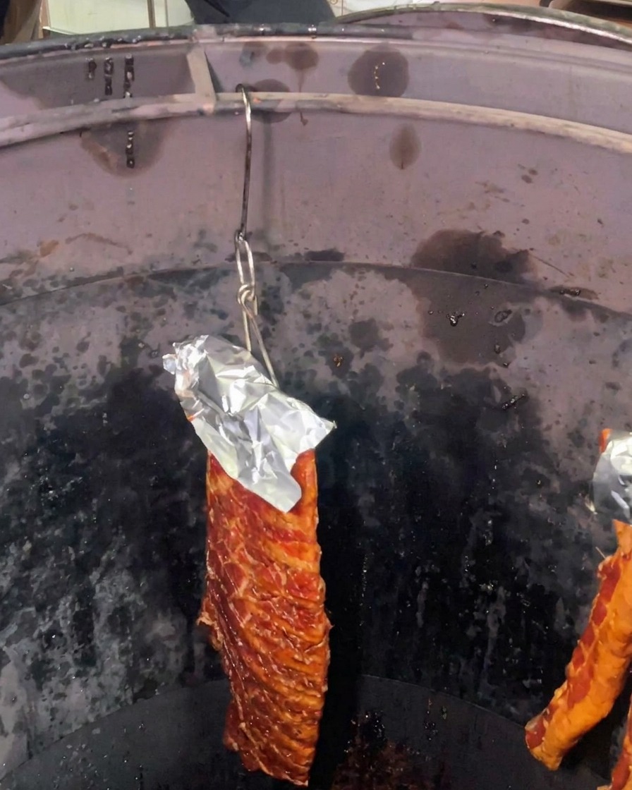Un costillar de costillas de cerdo marinadas con papel de aluminio protegiendo la parte superior colgando dentro de un horno de asado cilíndrico profundo y oscuro.