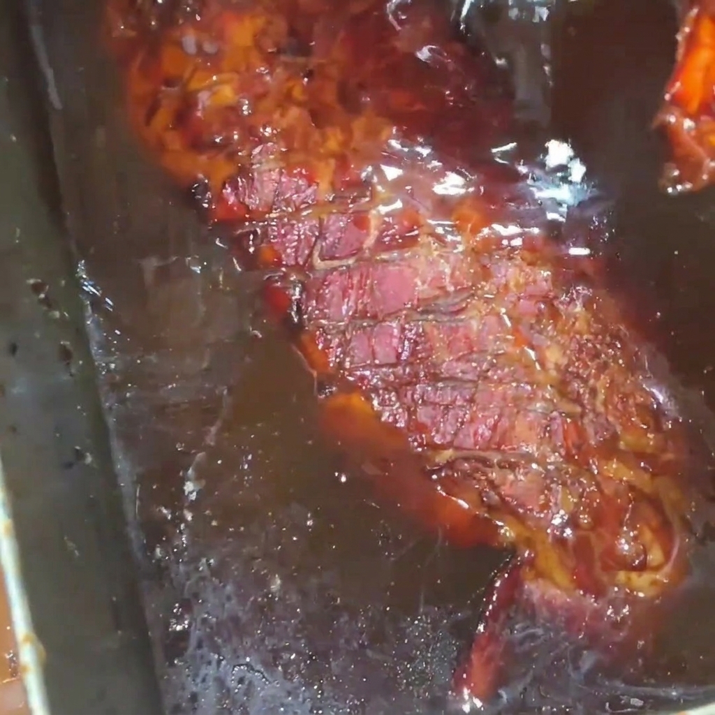 Un costillar de cerdo asado de color marrón dorado siendo bañado en un glaseado de jarabe de miel rico y brillante dentro de una bandeja de acero inoxidable.