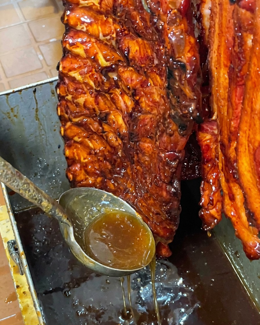 Un reluciente costillar de cerdo glaseado con miel colgando verticalmente sobre un recipiente de metal mientras el exceso de glaseado dulce gotea.