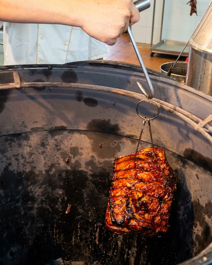 Una pieza de costillas de cerdo relucientes siendo bajada con un gancho de metal a un horno de asado cilíndrico profundo y oscuro.