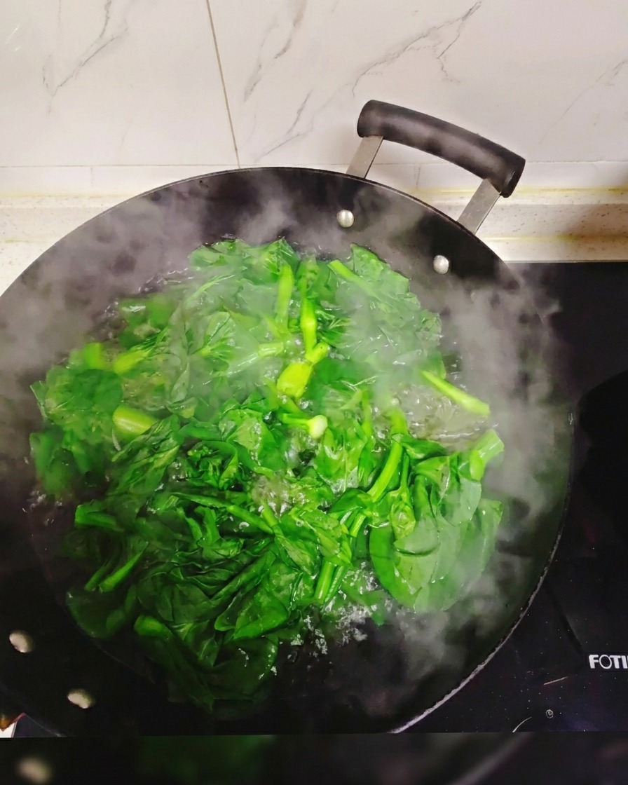 Verduras frescas de hoja verde sumergidas y blanqueándose en un wok grande lleno de agua hirviendo y humeante.