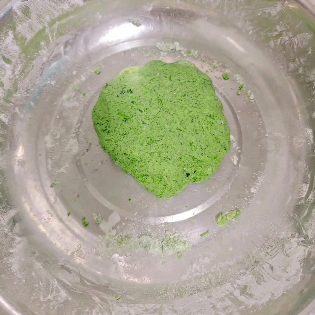 Una bola de masa de verduras suave y de color verde vibrante reposando dentro de un tazón de acero inoxidable con restos de harina en los bordes.