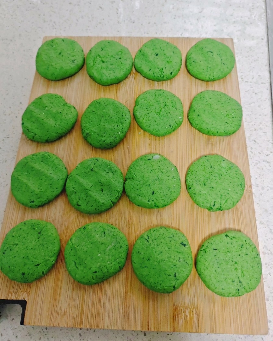 Dieciséis tortitas redondas de verdura verde dispuestas en un patrón de cuadrícula ordenada sobre una tabla de cortar de madera.