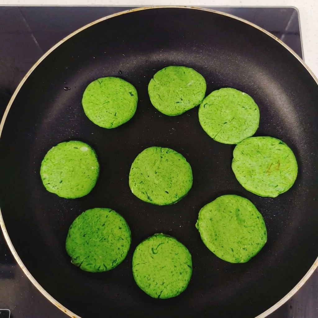 Varias tortitas redondas de color verde dispuestas en círculo dentro de una sartén antiadherente negra antes de cocinar.