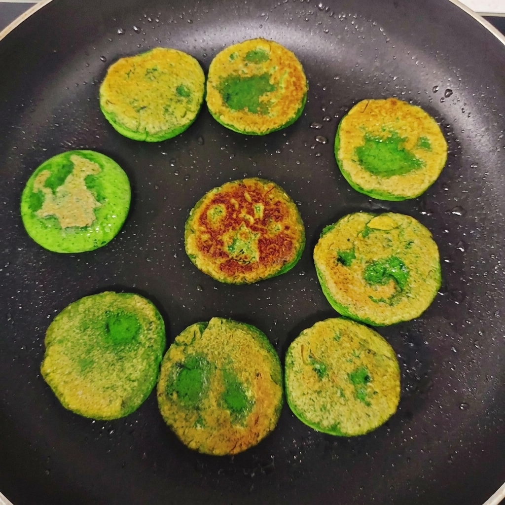 Tortitas de verdura verde volteadas en una sartén, mostrando costras doradas e irregulares en la parte superior.