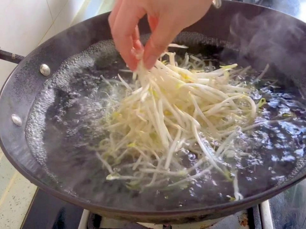 Un puñado de brotes de soja blancos y frescos cayendo en un wok con agua hirviendo.