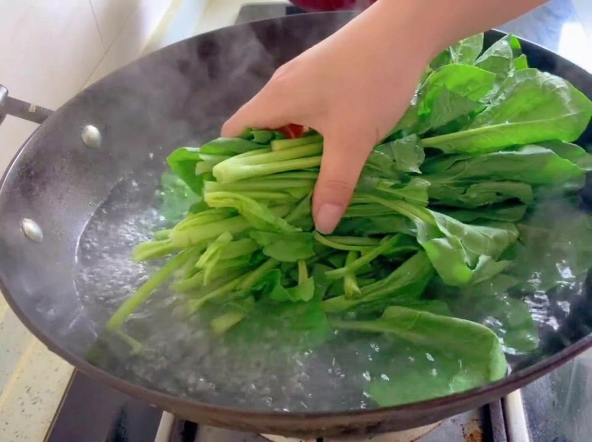 Un manojo de hojas de espinaca verdes y frescas introduciéndose en agua hirviendo dentro de un wok.