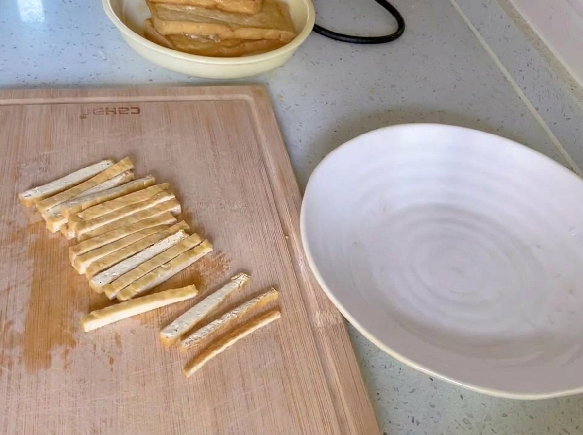 Tofu frito dorado cortado en tiras finas y uniformes sobre una tabla de cortar de madera junto a un plato blanco.