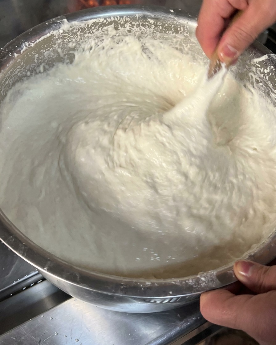 Primer plano de una masa blanca, espesa y pegajosa siendo levantada y estirada con un utensilio de madera dentro de un bol metálico.