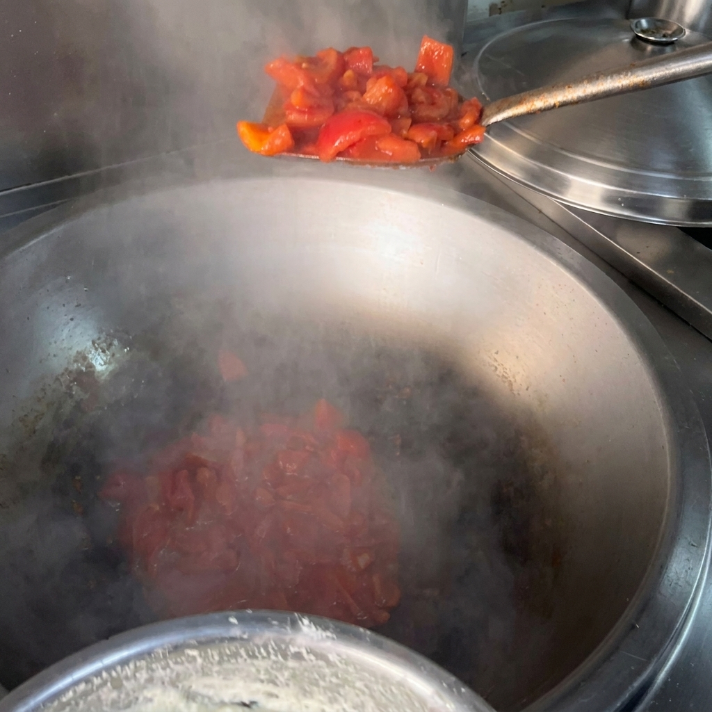 Una espátula de metal añadiendo tomates rojos picados a un wok de metal grande y humeante con aceite caliente.