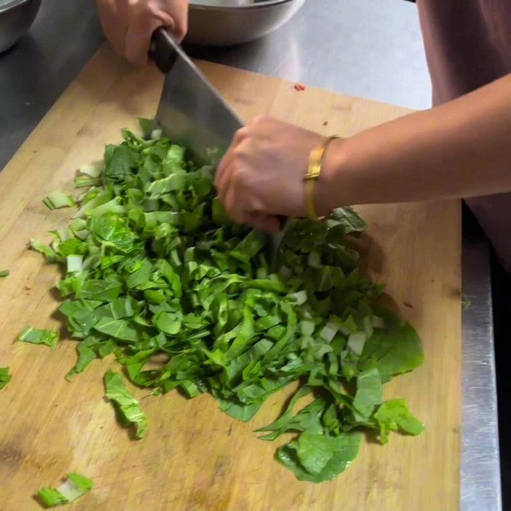 Manos usando un gran cuchillo de carnicero para picar verduras de hoja verde frescas sobre una tabla de cortar de madera.