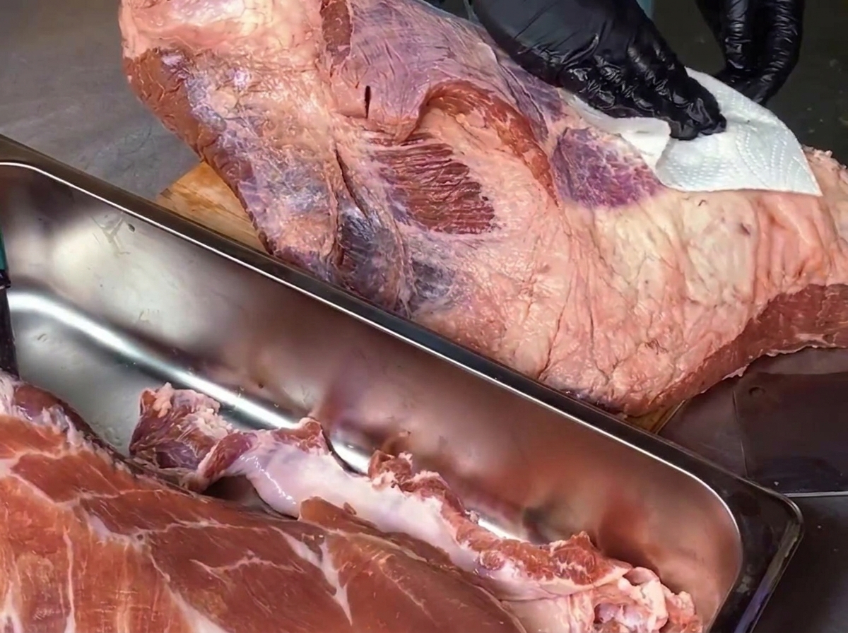 Un chef con guantes negros usa una toalla de papel blanca para secar un trozo grande de brisket de res crudo sobre una tabla de cortar de madera.