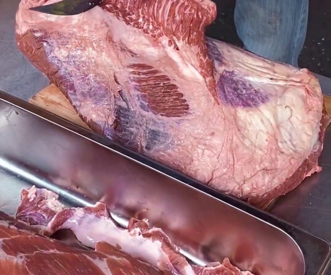 Primer plano de un chef utilizando un cuchillo afilado de forma paralela a la tabla de cortar para separar el músculo point del flat a lo largo de una línea de grasa blanca.