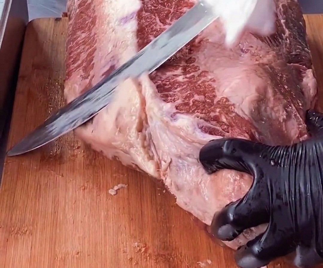 Una mano con guante sostiene una pieza de brisket mientras un cuchillo largo recorta una capa gruesa de grasa blanca de la carne roja.