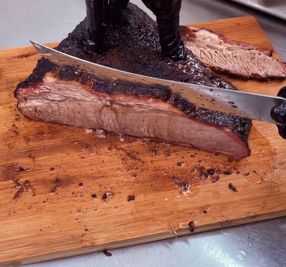 Un chef rebana un brisket de res bellamente ahumado con una costra oscura en sentido contrario a la fibra sobre una tabla de cortar de madera.