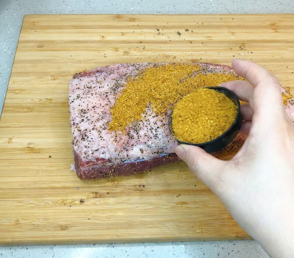 Mano sosteniendo un pequeño tazón negro y vertiendo un rub de barbacoa dorado sobre una gran pieza de brisket crudo en una tabla de madera.