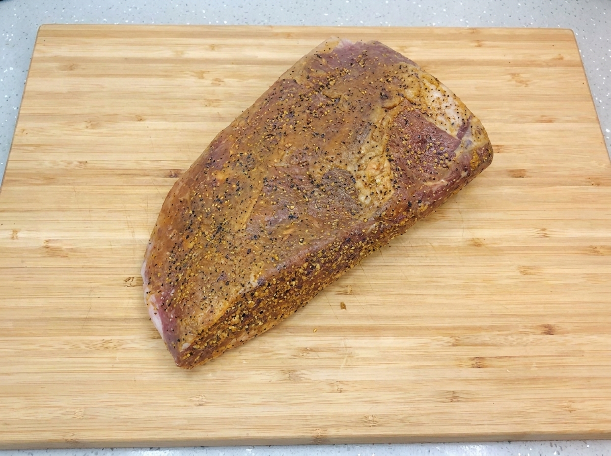 Brisket marinado reposando sobre una tabla de cortar de madera clara, brillante por una capa fresca de aceite y pimienta negra.