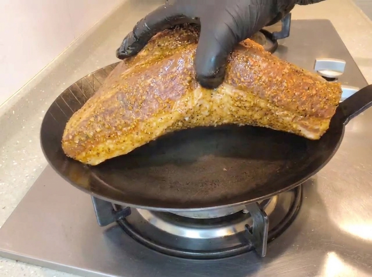 Mano con guante negro colocando un gran brisket sazonado con la grasa hacia abajo en una sartén de hierro caliente.