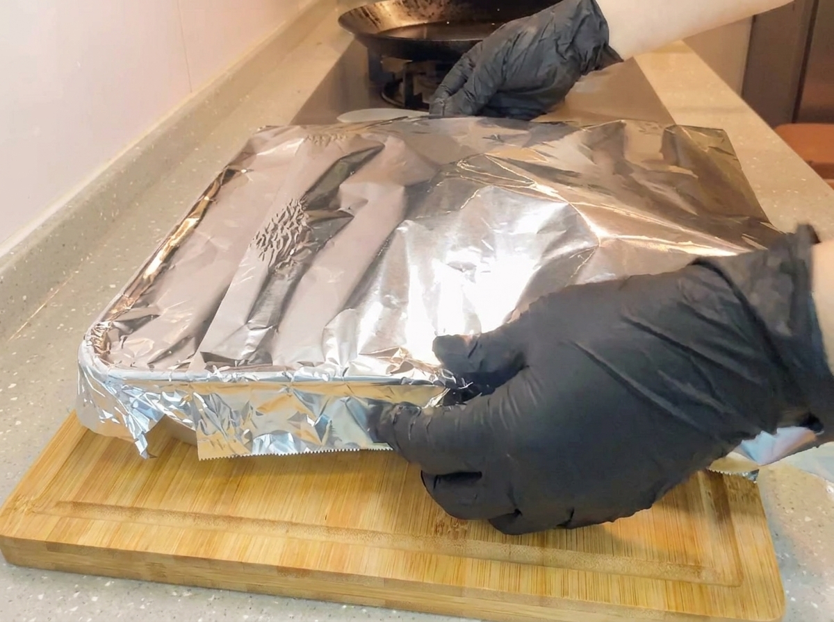 Manos con guantes envolviendo una nueva hoja de papel aluminio ajustadamente sobre un molde para hornear cuadrado en una tabla de madera.