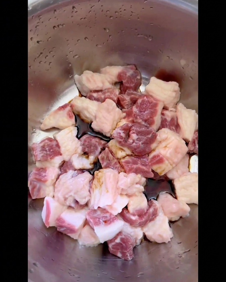 Salsa de soja oscura y condimentos líquidos cubriendo la grasa cruda de res en cubos dentro de un tazón de acero inoxidable.
