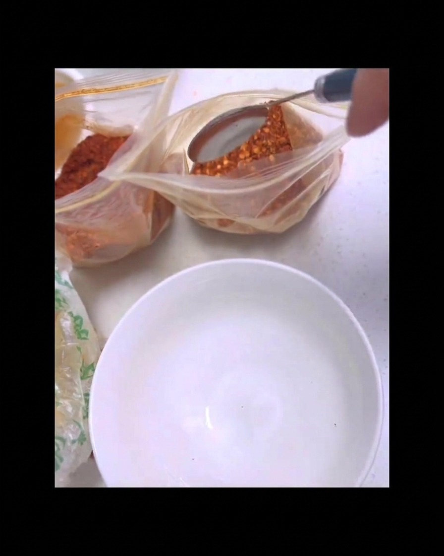 Una cuchara de metal sacando chile en polvo rojo de una bolsa de plástico transparente sobre un tazón blanco limpio.