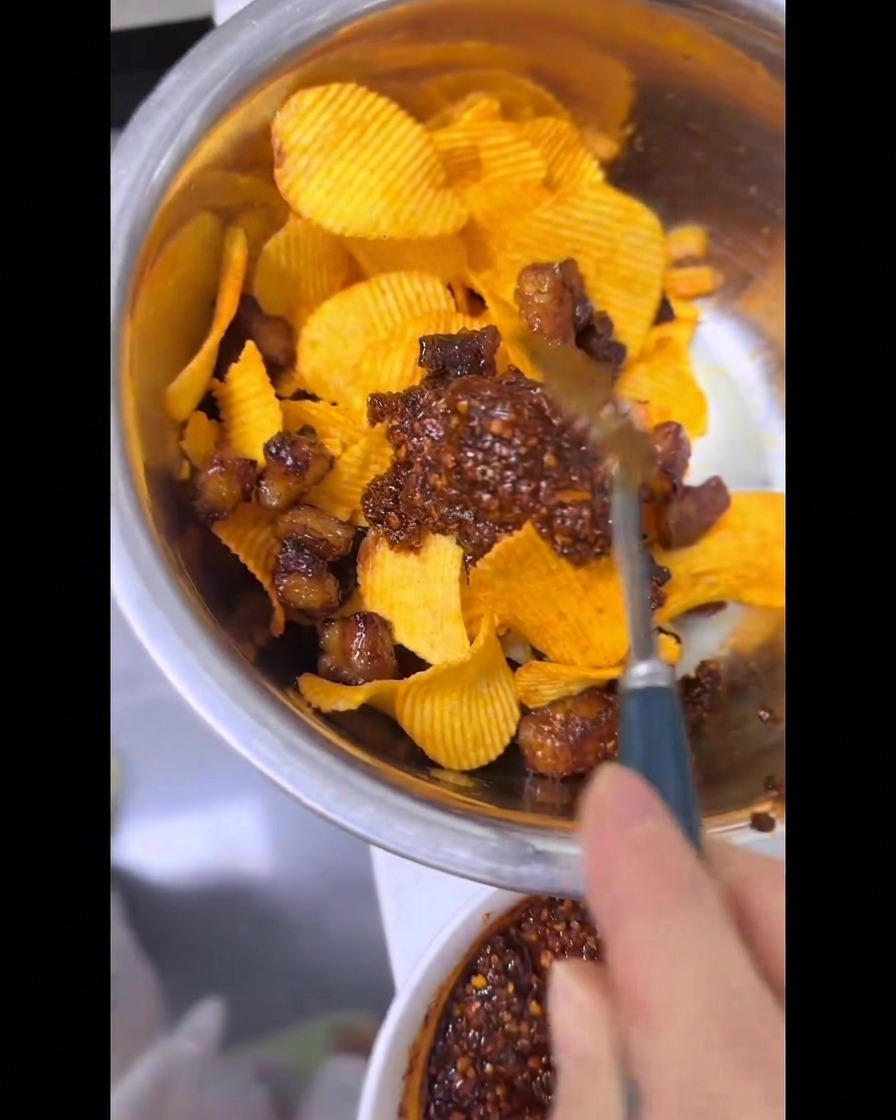 Una mano usando una cuchara para mezclar minuciosamente papas fritas onduladas, grasa de res crujiente y aceite de chile rojo brillante en un tazón de metal.