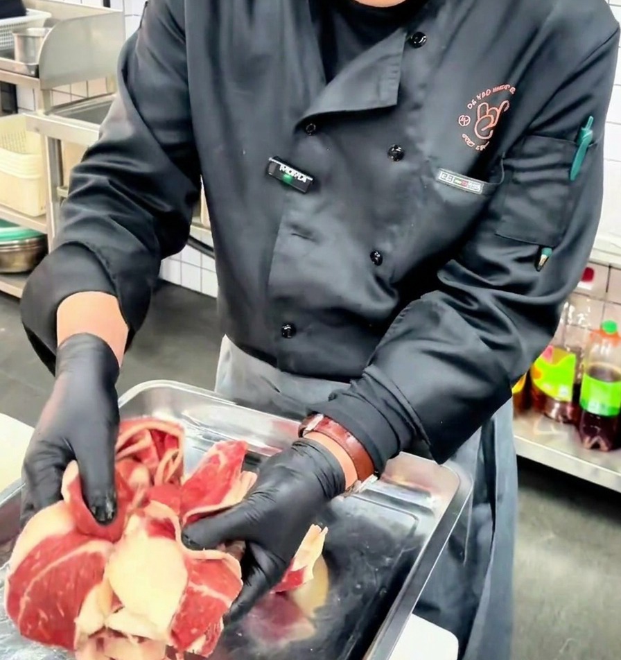 Un cocinero con guantes negros sosteniendo láminas finas de pecho de ternera cruda con capas visibles de grasa y carne sobre una bandeja metálica.