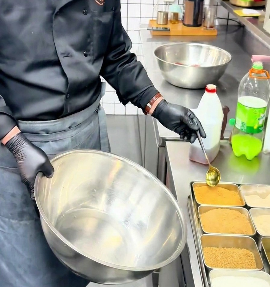 Un cocinero usando una cuchara dorada para añadir sazonador de carne en polvo en un bol grande de acero inoxidable.