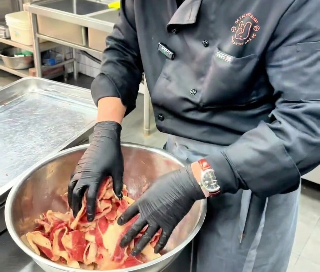 Un cocinero con guantes negros mezclando y masajeando la carne de ternera en láminas con el marinado dentro de un bol de acero inoxidable.