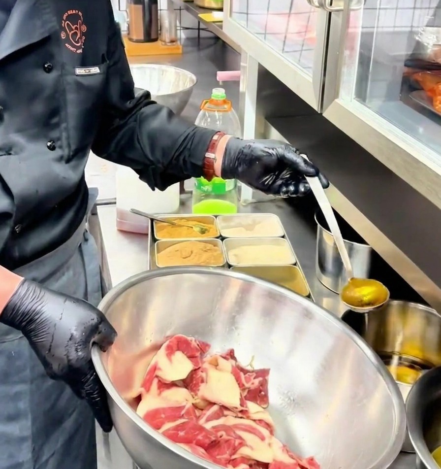 Un cucharón vertiendo aceite aromatizado dorado sobre las láminas de carne marinadas en un bol de acero inoxidable.