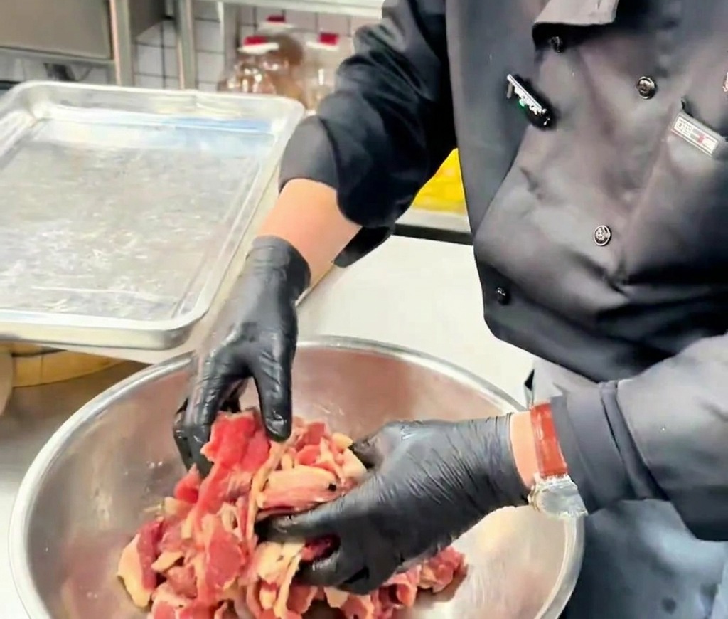 Manos con guantes mezclando láminas finas de carne con aceite dentro de un bol metálico.