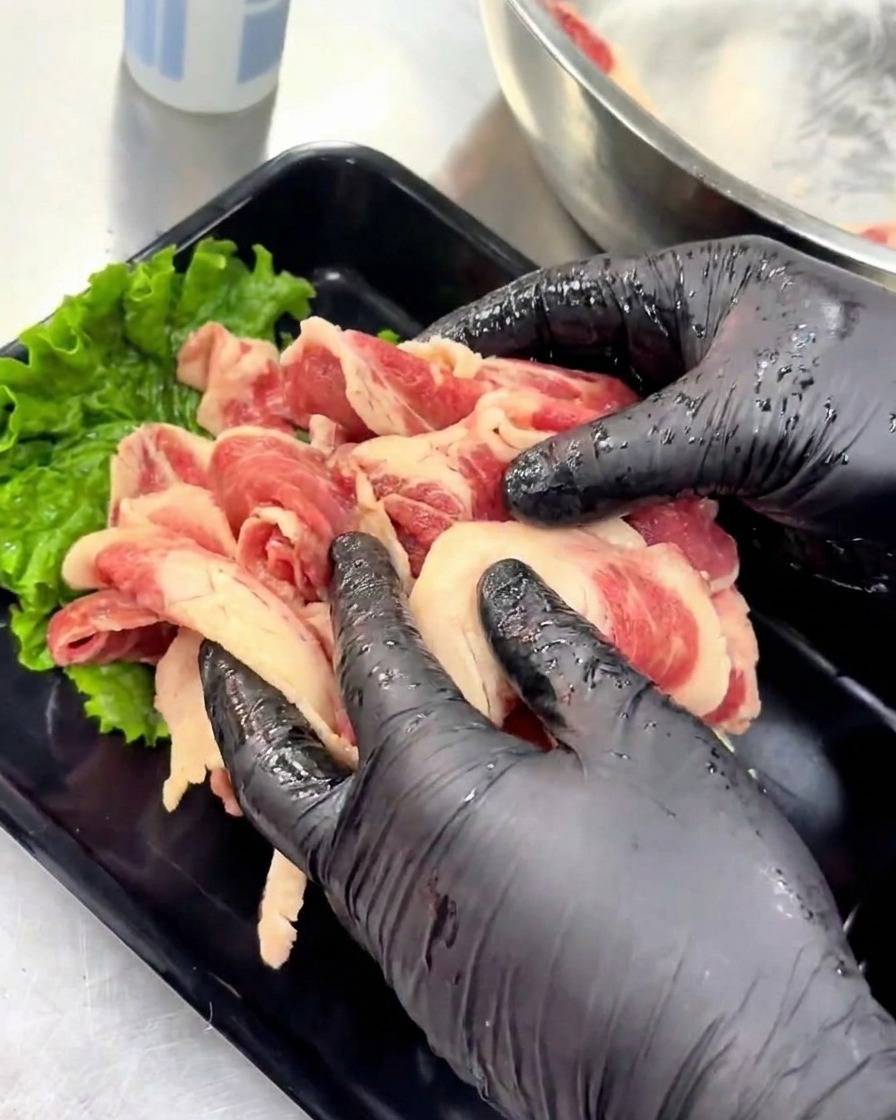 Manos con guantes acomodando ordenadamente las láminas de carne marinada cruda en una bandeja negra decorada con una hoja de lechuga verde.