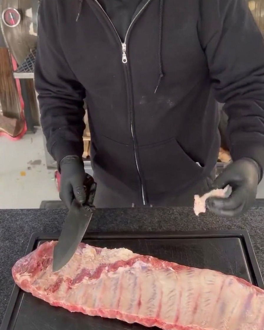 Una persona con guantes negros recorta el exceso de carne y los bordes irregulares de un costillar de cerdo crudo con un cuchillo de chef sobre una tabla de cortar negra.