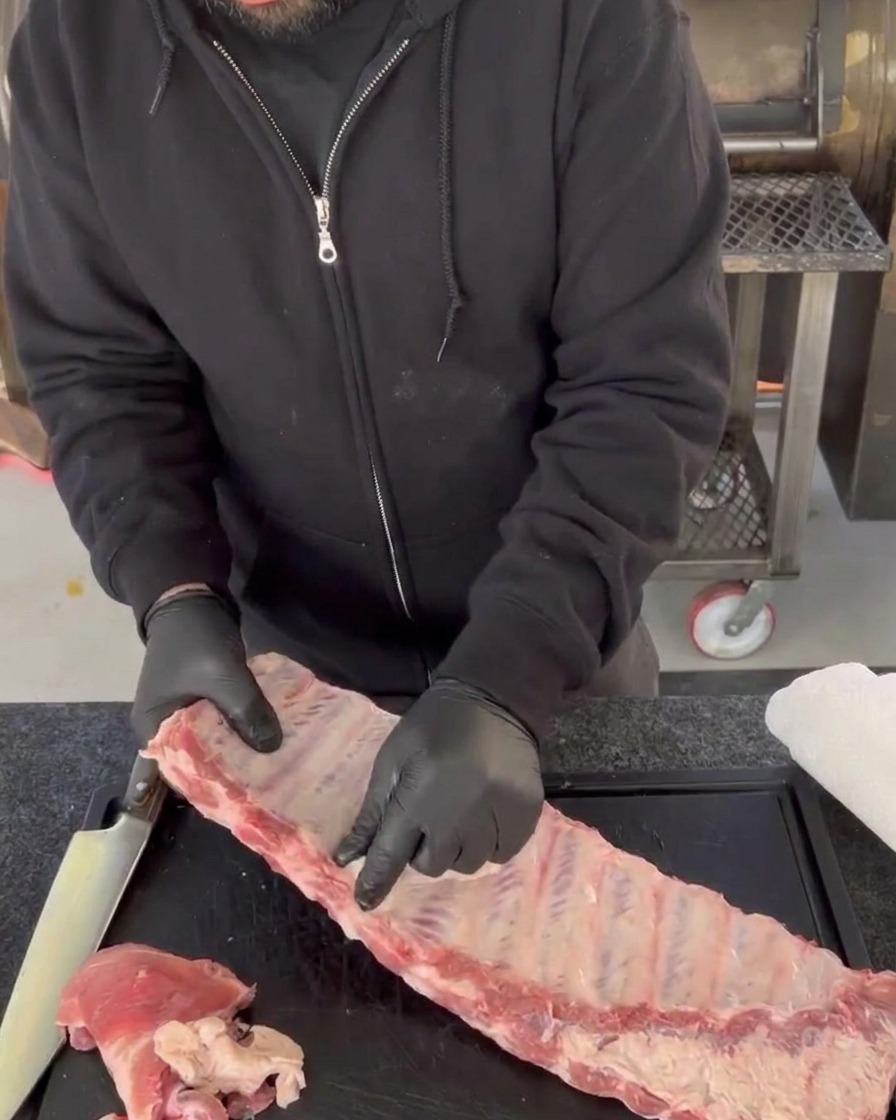 Manos con guantes despegando la membrana brillante y translúcida de los huesos posteriores de un costillar de cerdo crudo sobre una tabla de cortar.