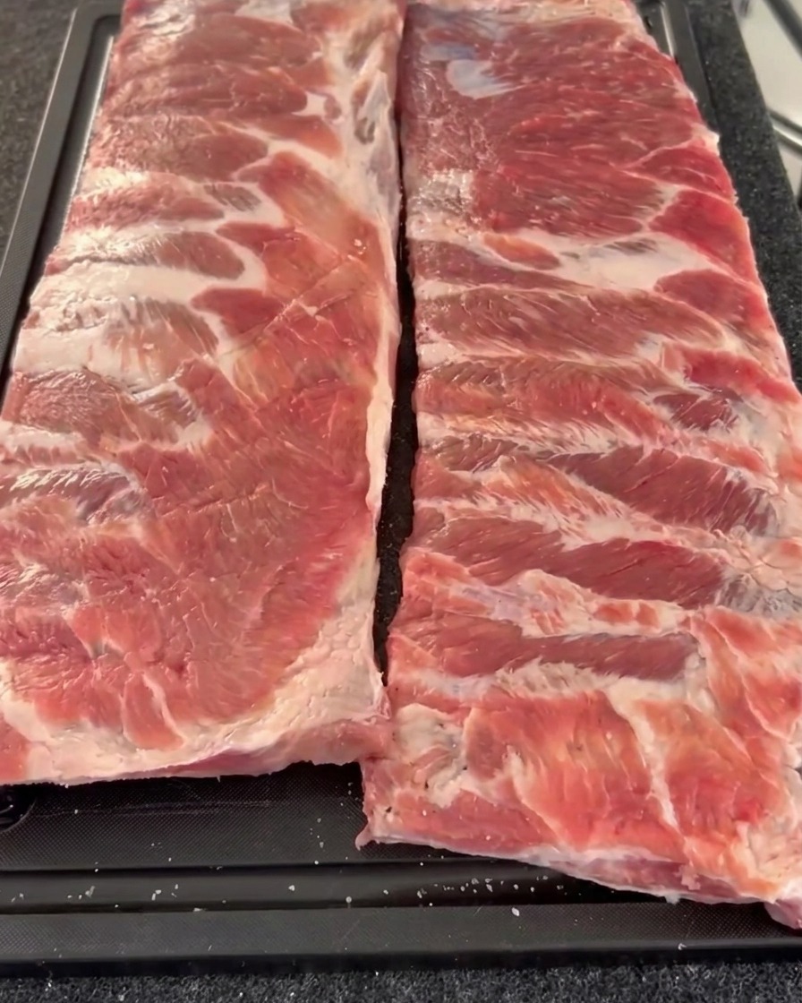 Dos costillares de cerdo crudos dispuestos uno al lado del otro en una tabla de cortar, mostrando el lado de la carne sazonado y cubierto con pimienta gruesa y sal.
